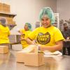 A teenage girl in a Cookie Cart shirt smiles at the camera as she packages cookies for deliver