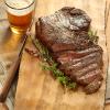 Cooked steak sliced on a wooden cutting board next to a tall glass of beer